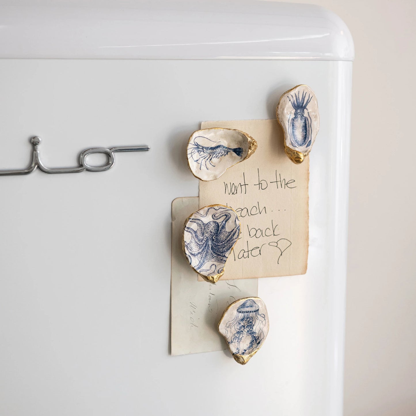 Set of four resin oyster shell-shaped magnets with sea life illustrations in white and blue, accented with gold, displayed on a metal card.