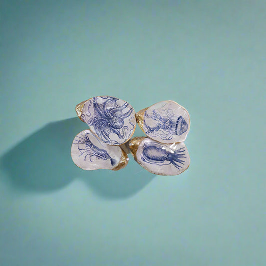 Set of four resin oyster shell-shaped magnets with sea life illustrations in white and blue, accented with gold, displayed on a metal card.